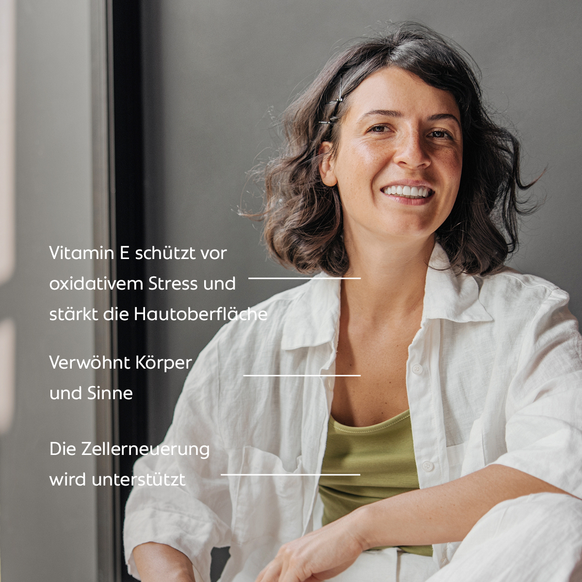 Eine lächelnde Frau sitzt entspannt am Fenster; der Text beschreibt, dass Vitamin E vor oxidativem Stress schützt, die Hautoberfläche stärkt, Körper und Sinne verwöhnt und die Zellerneuerung unterstützt.