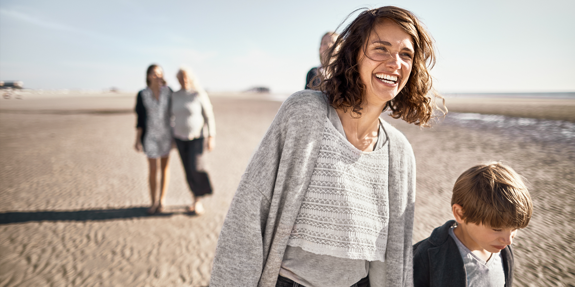 Frau mit lockigen braunen Haaren und Kind bei einem Spaziergang am Strand