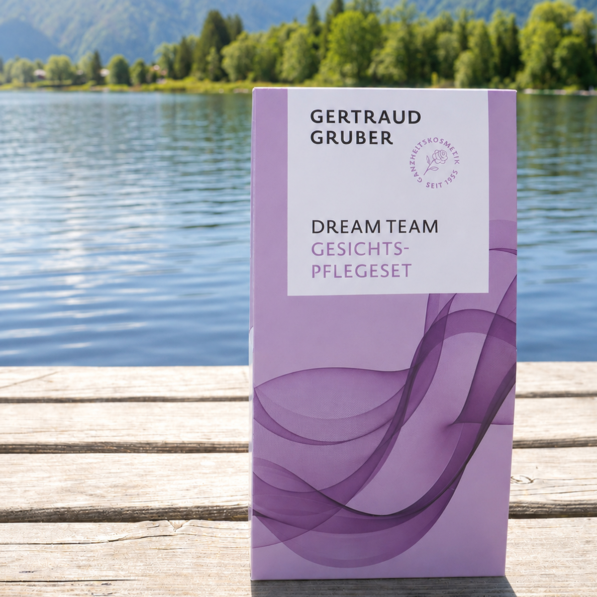 Lila Verpackung des Gertraud Gruber Dream Team Gesichtspflegesets auf einem Holzsteg vor einem See mit Bäumen und Bergen im Hintergrund.