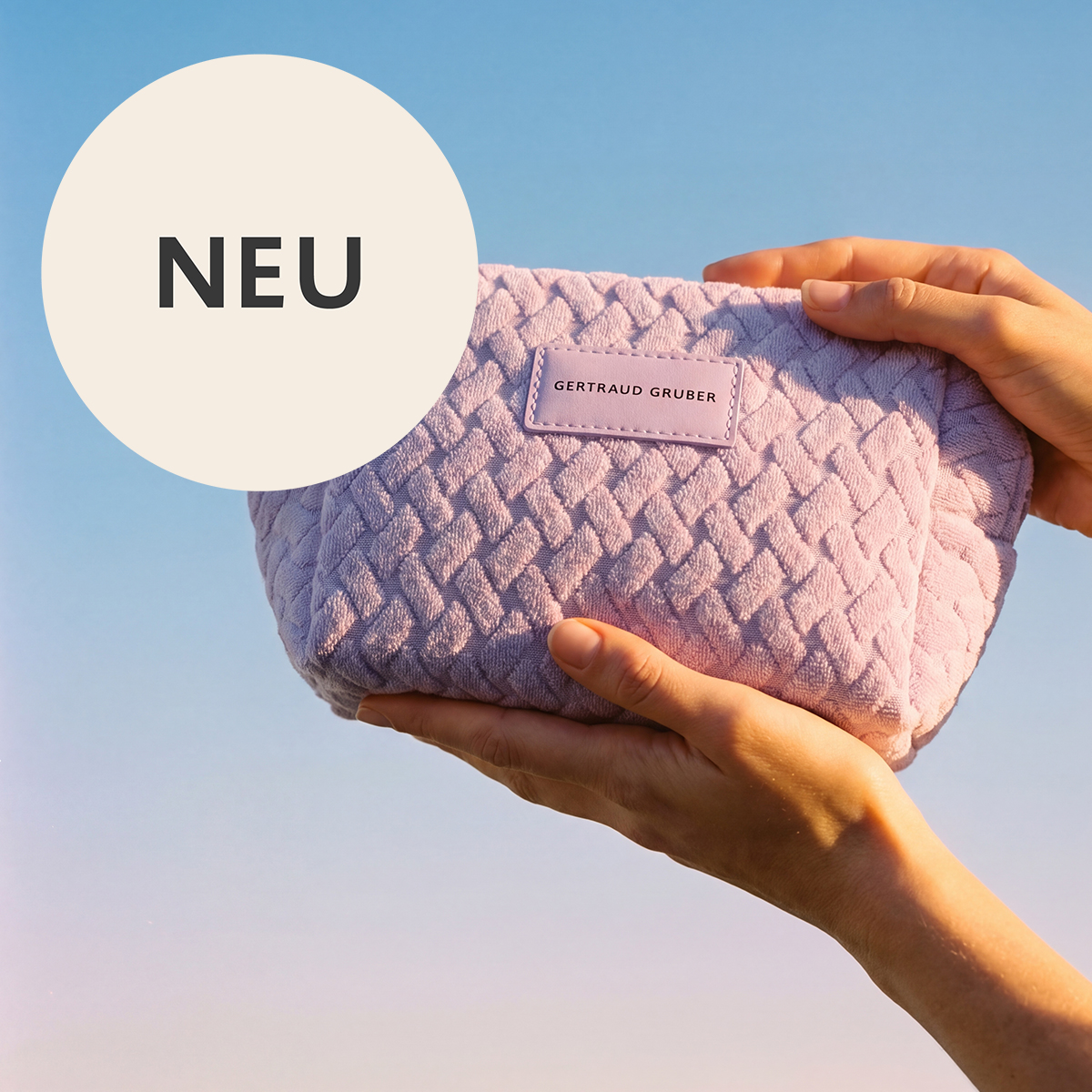 Zwei Hände halten eine strukturierte, hellviolette Kosmetiktasche vor einem klaren blauen Himmel. Die Tasche trägt ein Etikett mit der Aufschrift "Gertraud Gruber".