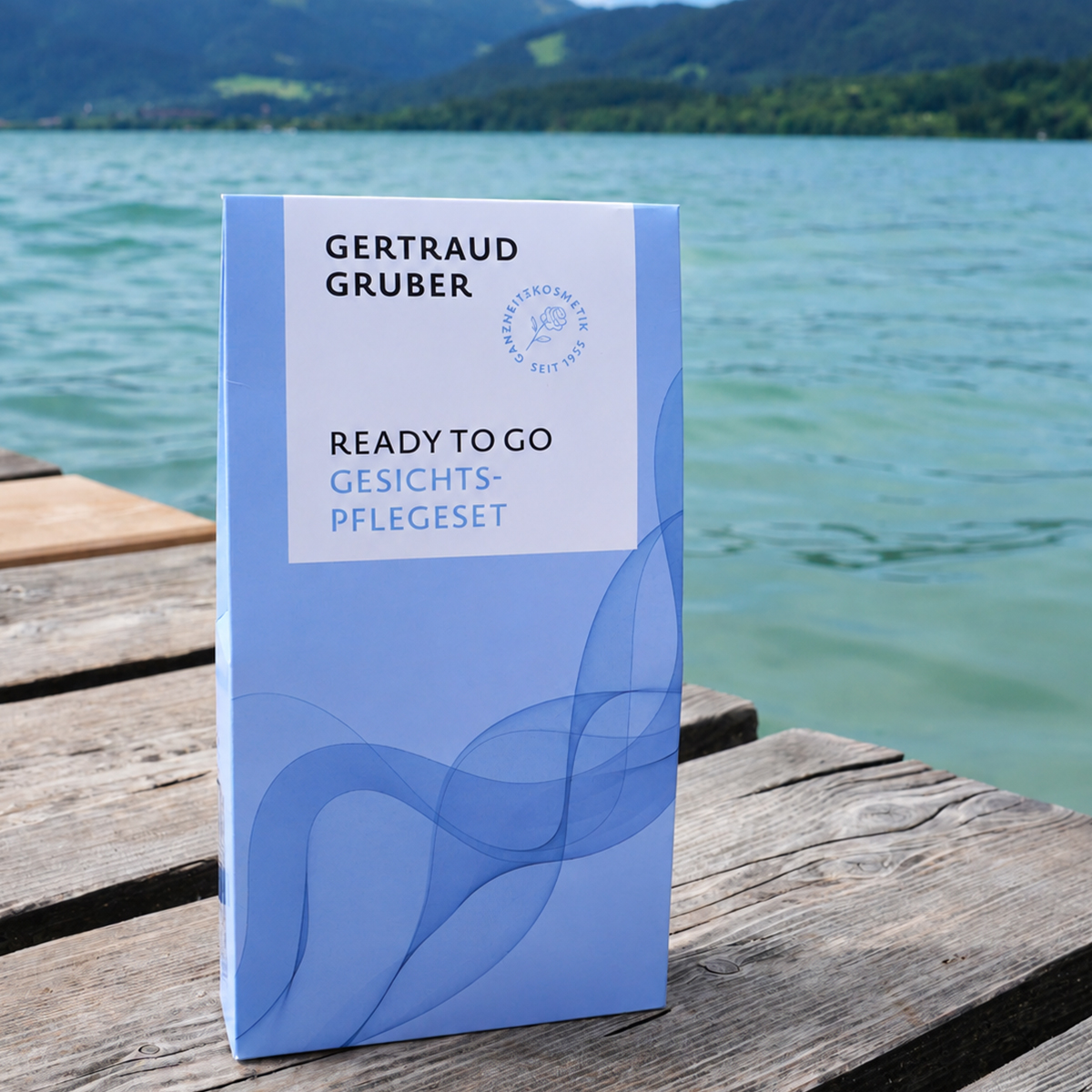 Blaue Verpackung des Gertraud Gruber Ready to Go Gesichtspflegesets auf einem Holzsteg vor einem See mit Bergen im Hintergrund.