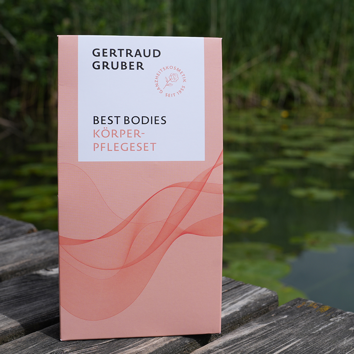 Rosafarbene Verpackung des Gertraud Gruber Best Bodies Körperpflegesets auf einem Holzsteg vor einem Teich mit Schilf im Hintergrund.